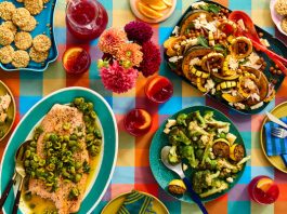 A colorful tablecloth is topped with platters of food, drink, plates and flowers.