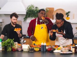Daniel Radcliffe, Tracy Morgan and Erika Alexander stand at a kitchen counter eating pizza.