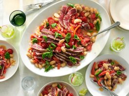 A white platter with seared tuna, cooked beans, tomatoes, peppers and herbs is shown on a table alongside a light green cucumber drink garnished with a sliced radish.