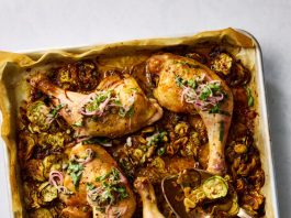 Roasted chicken legs topped with sliced red onions and herbs rest on a baking sheet layered with cooked zucchini.