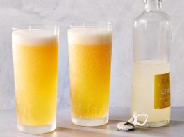 It’s Not Summer Until You’ve Had a Proper Shandy Frosted glasses hold a sparkling shandy with foam. A glass bottle with a yellow label and a bottle opener are nearby.