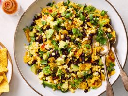 A vibrant salad in a large white bowl featuring corn, black beans, avocado chunks and crumbled tortilla chips, garnished with fresh cilantro. A bottle of orange hot sauce and a plate of golden tortilla chips are in the background.