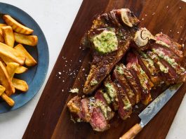 A steak topped with herb butter, sliced on a cutting board next to a bowl full of French fries.