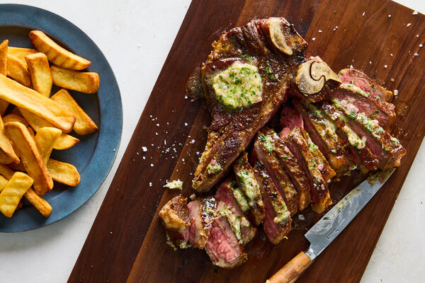 A steak topped with herb butter, sliced on a cutting board next to a bowl full of French fries.