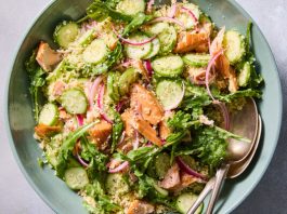 A teal bowl of mixed salad with couscous, salmon, sliced cucumbers, red onions and arugula. Two spoons are visible inside.