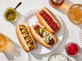 An overhead image of three hot dogs on a plate surrounded by drinks.