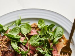 A white plate holds fanned slices of steak, garnished with a chopped brown relish and bright green leaves. A golden fork is on the right side.