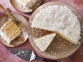 A coconut cake on a speckled stand with a missing slice, revealing its white cake and bright yellow filling. A cake server and a plate with a slice are also shown.