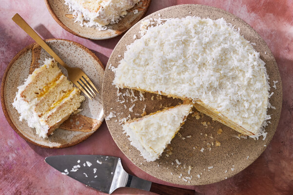 A coconut cake on a speckled stand with a missing slice, revealing its white cake and bright yellow filling. A cake server and a plate with a slice are also shown.