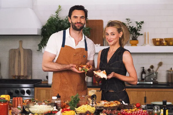 Tom Bateman with Lili Reinhart at a kitchen counter filled with pizza toppings.