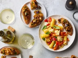 Let This Dinner Menu Take You to Sicily, No Passport Needed An overhead image of a party spread that includes a Dutch oven filled with pasta, surrounded by plates topped with caponata toasts.