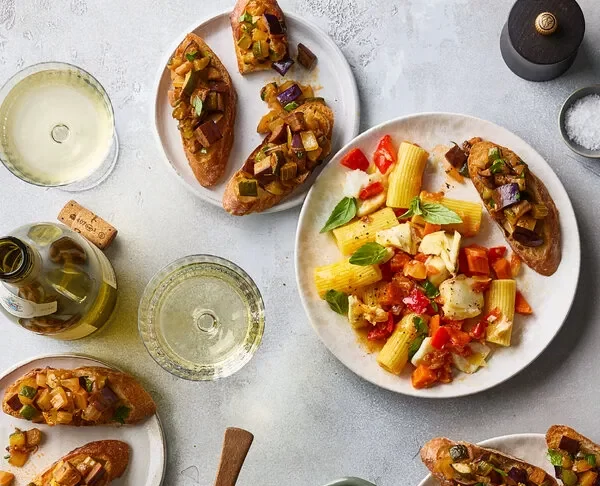 An overhead image of a party spread that includes a Dutch oven filled with pasta, surrounded by plates topped with caponata toasts.