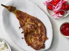 Why I Can’t Stop Making Eggplant Omelets, Filipino-Style An overhead image of a long piece of eggplant that’s coated with egg. A plate of onion and tomato, some ketchup and a bowl of rice surround.