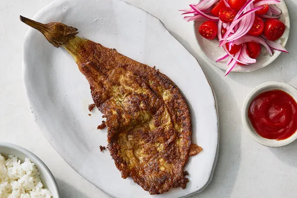 Why I Can’t Stop Making Eggplant Omelets, Filipino-Style An overhead image of a long piece of eggplant that’s coated with egg. A plate of onion and tomato, some ketchup and a bowl of rice surround.